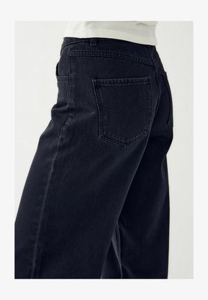 Prim-plan al unei persoane care poartă blugi negri din denim, arătând buzunarul din spate și cusătura laterală pe un fundal deschis.