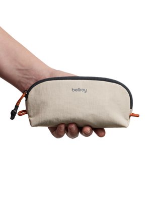 Mano che tiene una pochette Bellroy beige con bordatura della cerniera nera e tiretti della cerniera arancioni su sfondo bianco.