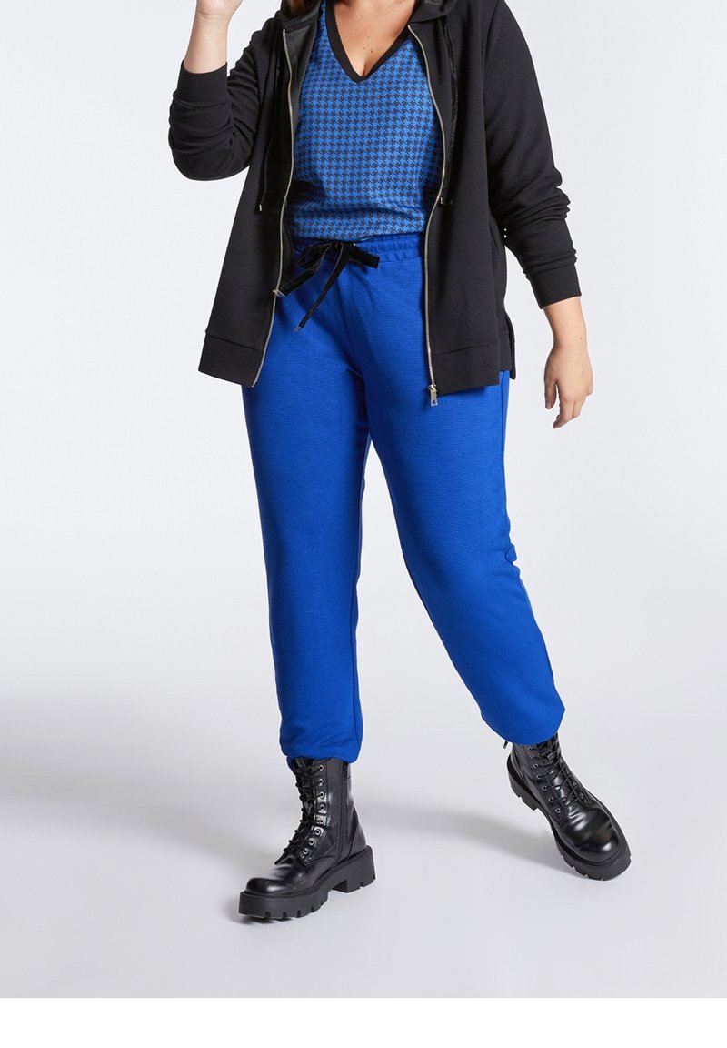 Modré tepláky s elastickým pasem a šňůrkou, spárované s modrým vrchním trikem s vzorem houndstooth a černou zipovou mikinou. Robustní černé boty.