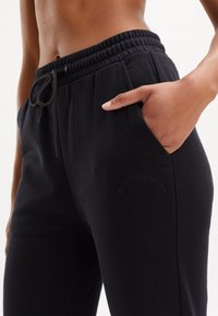 Black sweatpants with elastic waistband and drawstring, featuring side pockets and tonal embroidered logo on the thigh. Smooth cotton material.