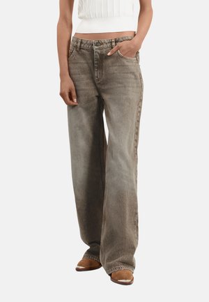 Vrouw met vervaagde bruine wijde jeans met hand in de zak, gecombineerd met een wit mouwloos topje en bruine schoenen.