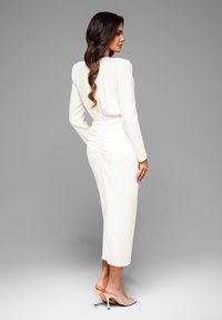 Weißes langärmeliges Kleid mit gerafftem Rückendesign, mittellanger Stil, kombiniert mit glitzernden High-Heels. Glattes, fließendes Material.