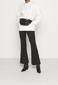 Kermakotelo, jossa on säädettävä naru, yhdistettynä mustiin levenneisiin housuihin ja mustaan vyölaukkuun. Mallilla on päällään mustat korkeat nilkkasaappaat.