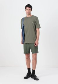 T-shirt en coton vert olive avec un logo, associé à un short assorti. Tient un sac en denim bleu. Des chaussures et des chaussettes noires complètent le look.