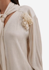 Crème-kleurige blouse met een textuurstof, voorzien van een gestrikt decolleté en een kanten bloemenaccent op de linkerschouder.