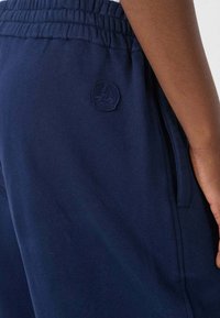 Pantalones de chándal azul marino con cintura elástica, bolsillos laterales y un parche de logo texturizado en el muslo izquierdo. Material de algodón suave.