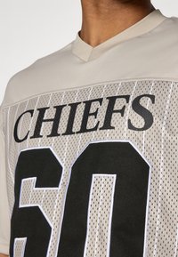 Μπεζ δικτυωτό μπούστο με μαύρο κείμενο "CHIEFS" και μεγάλο αριθμό "60". Έχει αθλητικό σχέδιο και κοντό γιακά.