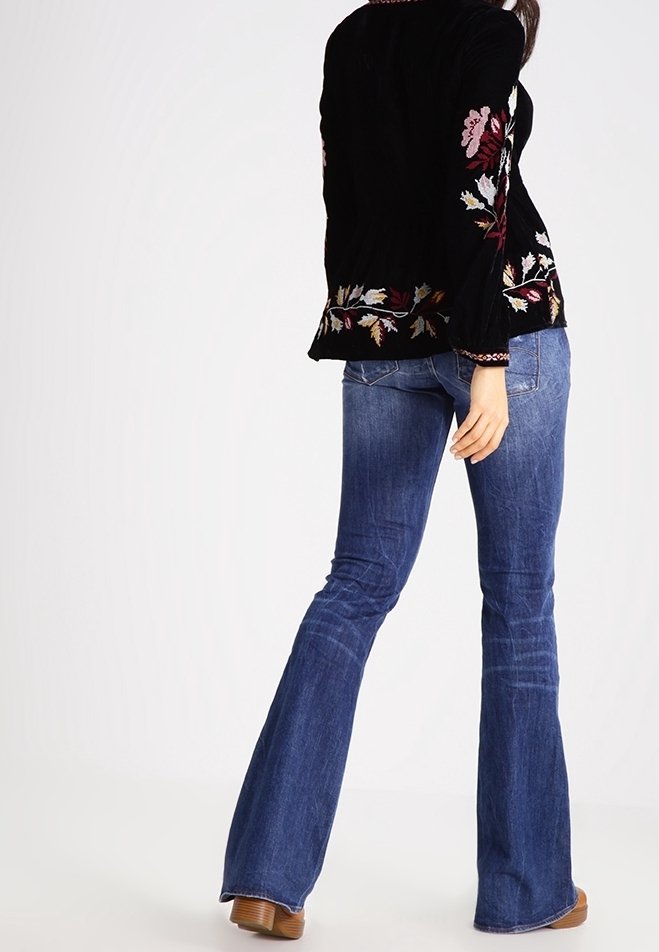 Veste en velours côtelé noire avec broderies florales colorées, assortie à un jean bleu évasé. La tenue est complétée par des chaussures beiges.