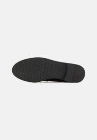 Semelle de chaussure noire avec une surface texturée présentant un motif de logo répétitif, des bords lisses et un léger talon pour une meilleure adhérence et stabilité.