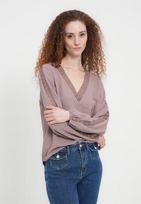 Μάβ χαλαρή μπλούζα με βαθύ V-λαιμόκοψη, περίτεχνη δαντέλα στα μανίκια και απαλό, υφασμάτινο ύφασμα συνδυασμένο με μπλε τζιν.