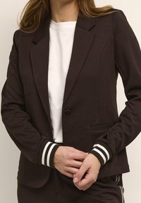 Nainen, jolla on yllään tummanruskea bleiseri, jossa on raidalliset hihansuut, valkoisen paidan päällä, kädet ristissä vyötärön ympärillä neutraalissa taustassa.