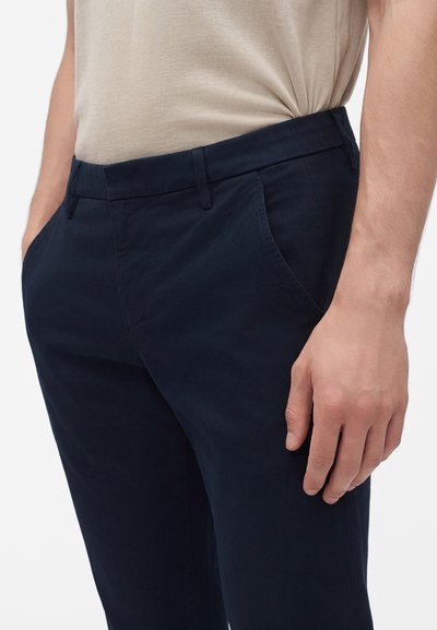 Homme portant un pantalon bleu marine avec des poches latérales et une chemise beige rentrée, debout avec une main détendue sur le côté.