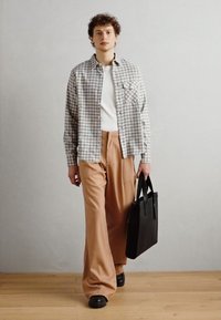 Modelo vistiendo una camisa a cuadros de color gris claro y blanco, sobre una camiseta blanca, combinada con pantalones beige holgados y sosteniendo una bolsa tote negra.