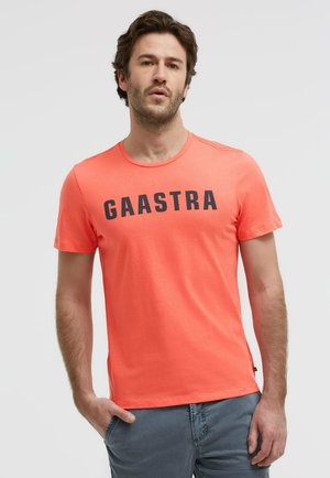 Mann trägt ein korallenfarbenes T-Shirt mit kurzen Ärmeln und dem Schriftzug "GAASTRA" sowie graue Hosen, steht mit einer Hand in der Tasche vor einem einfarbigen Hintergrund.