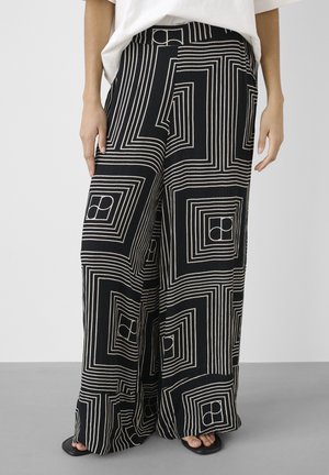 Osoba nosząca czarne spodnie o szerokich nogawkach z białymi geometrycznymi wzorami kwadratów i kół, zestawione z czarnymi sandałami na otwartym palcu oraz białą bluzką.