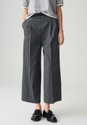 Person trägt graue Nadelstreifen-Hosen mit weitem Bein, hellgraues kurzärmeliges Oberteil und schwarze Loafers, steht mit einer Hand in der Tasche.