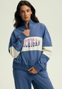 Niebieska bluza z suwakiem z kremowym panelem środkowym i napisem "MICHIGAN" w kolorze różowym. Dzianinowe mankiety i pas; połączona ze spodniami w tym samym kolorze.