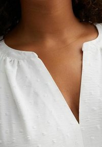 Detailní záběr na krk a horní část hrudníku osoby v texturované bílé blůze s výstřihem ve tvaru V a nařasenými detaily látky.