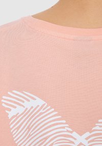 Camiseta de algodón rosa claro con cuello redondo, que presenta un patrón abstracto de hojas blancas en el centro. Textura suave con mangas cortas.