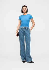 Γυναίκα που στέκεται φορώντας εφαρμοστή μπλε μπλούζα με κοντά μανίκια, μπλε παντελόνι με φαρδιά παντελόνια, μαύρα μυτερά παπούτσια και κρατώντας μια μαύρη τσάντα χειρός.