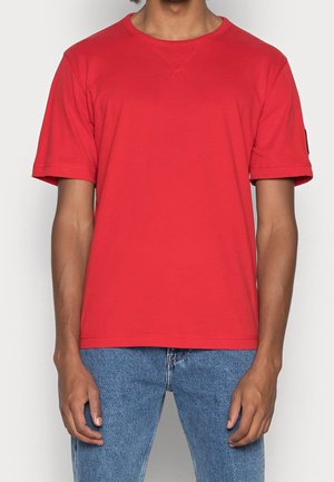 Punainen puuvillainen t-paita, jossa on lyhyet hihat, pyöreä pääntie ja rento istuvuus. Näkyvät ompelusaumat ja logopatch hihassa.