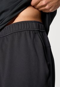 Μαύρο αθλητικό σορτς με ελαστική μέση και τσέπες, κατασκευασμένο από λείο, ελαστικό υλικό, που δείχνει τη μέση πάνω από ένα εφαρμοστό τοπ.