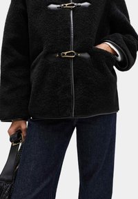 Giacca in pile nera con zip frontale, caratterizzata da accessori dorati e dettagli in pelle. Ha una vestibilità comoda e tasche laterali.