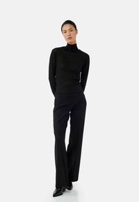 Pull en col roulé noir ajusté avec un motif texturé, associé à un pantalon noir évasé taille haute. Modèle portant des chaussures à talons noires.