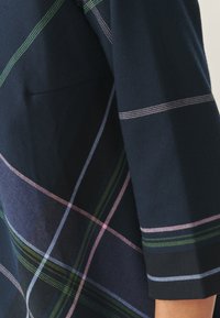 Marineblaue Stoff mit einemKaromuster in grünen, rosa und blauen Linien. Verfügt über ein einfaches Design und sichtbare Nahtdetails.