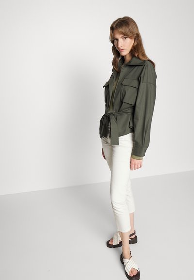 Camisa jaqueta verde-oliva de mangas longas com cintura marcada, apresentando dois bolsos no peito e um colarinho dobrado, emparelhada com calças brancas cropped e sandálias.