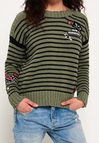 Maglione a righe verdi e nere lavorato a maglia con orlo a coste, caratterizzato da varie toppe grafiche, inclusi motivi di stelle e fulmini.