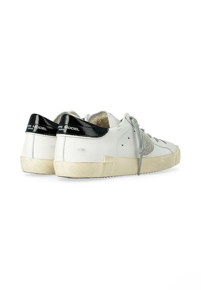 Baskets basses blanches avec des languettes de talon noires brillantes, des lacets gris, et des semelles beige légèrement usées.