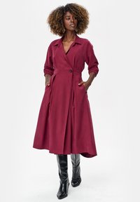 Kobieta o kręconych włosach w bordowej rozpinanej na guziki sukience midi z kieszeniami oraz czarnych kozakach za kolano, stojąca na jednolitym tle.