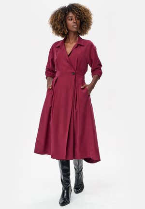 Kobieta o kręconych włosach w bordowej rozpinanej na guziki sukience midi z kieszeniami oraz czarnych kozakach za kolano, stojąca na jednolitym tle.