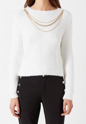 Woman wearing a white fuzzy long-sleeve sweater with layered gold chains and black pants featuring gold buttons on the sides.