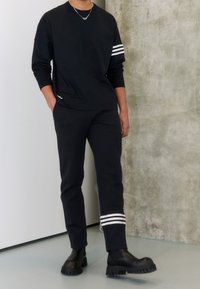 Černá košile s dlouhým rukávem se třemi bílými pruhy na rukávech, doplněná černými kalhotami se zúženými nohavicemi a robustními černými botami. Hladká textura látky.