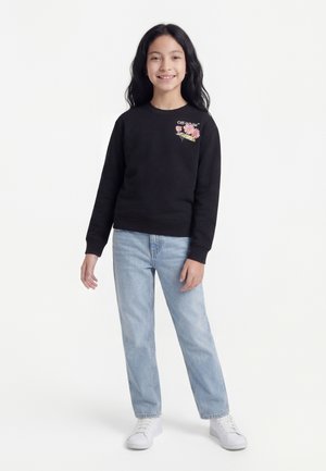 Pige med langt sort hår iført sort sweatshirt med blomsterdesign og lyseblå jeans, stående og smilende på hvid baggrund.