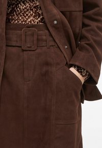Manteau en cuir suédine marron avec une ceinture assortie, présentant une couche intérieure texturée et une poche latérale. Boutons à pression visibles à l'avant.
