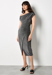 Femme portant une robe midi asymétrique à une épaule scintillante avec une fente à l'avant et des mules noires à talons ouverts.