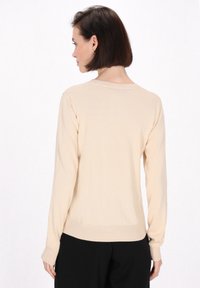 Jasno-beżowy sweter z długimi rękawami, ściągaczami na nadgarstkach i na dole. Miękka faktura, prosty design i okrągły dekolt. Czarna spodnie na pierwszym planie.