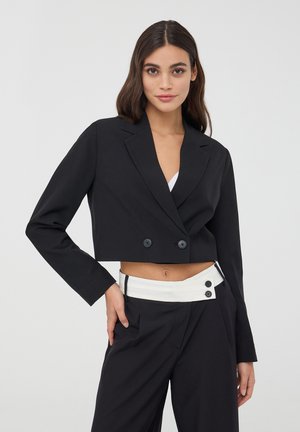 Blazer nero accostato con colletto a rever, due bottoni neri e maniche lunghe. Abbinato a pantaloni a vita alta con dettagli bianchi.