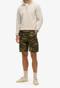 Chaqueta de forro polar beige con cremallera, pantalones cortos de camuflaje con múltiples bolsillos y zapatillas de color beig claro. Tejido suave y ajuste relajado.