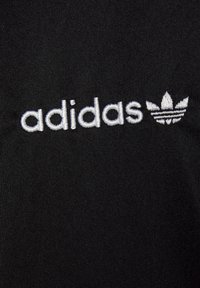 Tela negra con superficie texturizada que presenta el texto bordado en blanco "adidas" y un logo de trébol encima del texto.