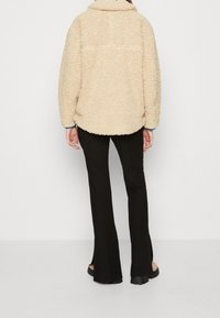 Béžová, huňatá fleecová bunda s vysokým límcem, oversized střih a kleslými rameny; nošena přes černé úzké kalhoty. Hladká textura bez vzorů.