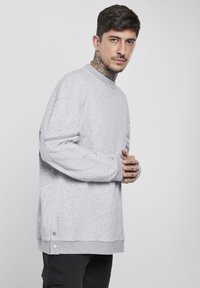 Sudadera gris con cuello redondo, bolsillo lateral con cremallera y detalles de snaps en el dobladillo. Tejido suave con un corte relajado y mangas largas.
