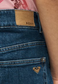 Σκούρο μπλε τζιν denim με ταμπά ραμμένη ετικέτα επωνυμίας Roxy και μικρό λογότυπο καρδιάς κοντά στην μπροστινή τσέπη, φορεμένα με ροζ φλοράλ τοπ.