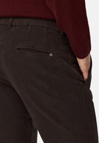 Tumši brūni kordūrveida bikses ar aizmugurējo kabatu, kam ir izteiksmīgs logo uzlīme. Audumam ir maiga tekstūra un smalka mirdzuma.