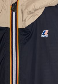 Giacca K-Way con una combinazione di colori beige e blu scuro. Presenta una cerniera frontale con strisce multicolori e un logo sul petto. Materiale leggero.