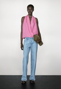 Roze gedrapeerde haltertop, lichtblauwe wijde jeans en donkere sandalen. Model houdt een bruine suède clutch vast. Eenvoudige, moderne styling.