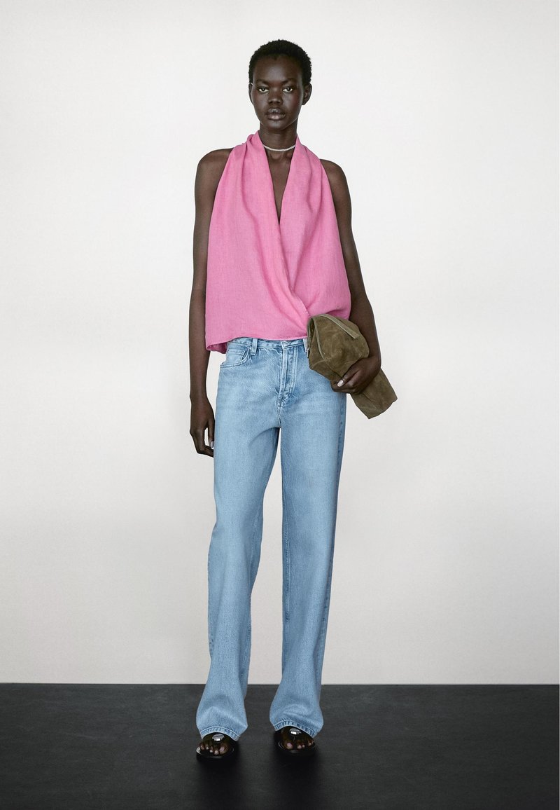Roze gedrapeerde haltertop, lichtblauwe wijde jeans en donkere sandalen. Model houdt een bruine suède clutch vast. Eenvoudige, moderne styling.